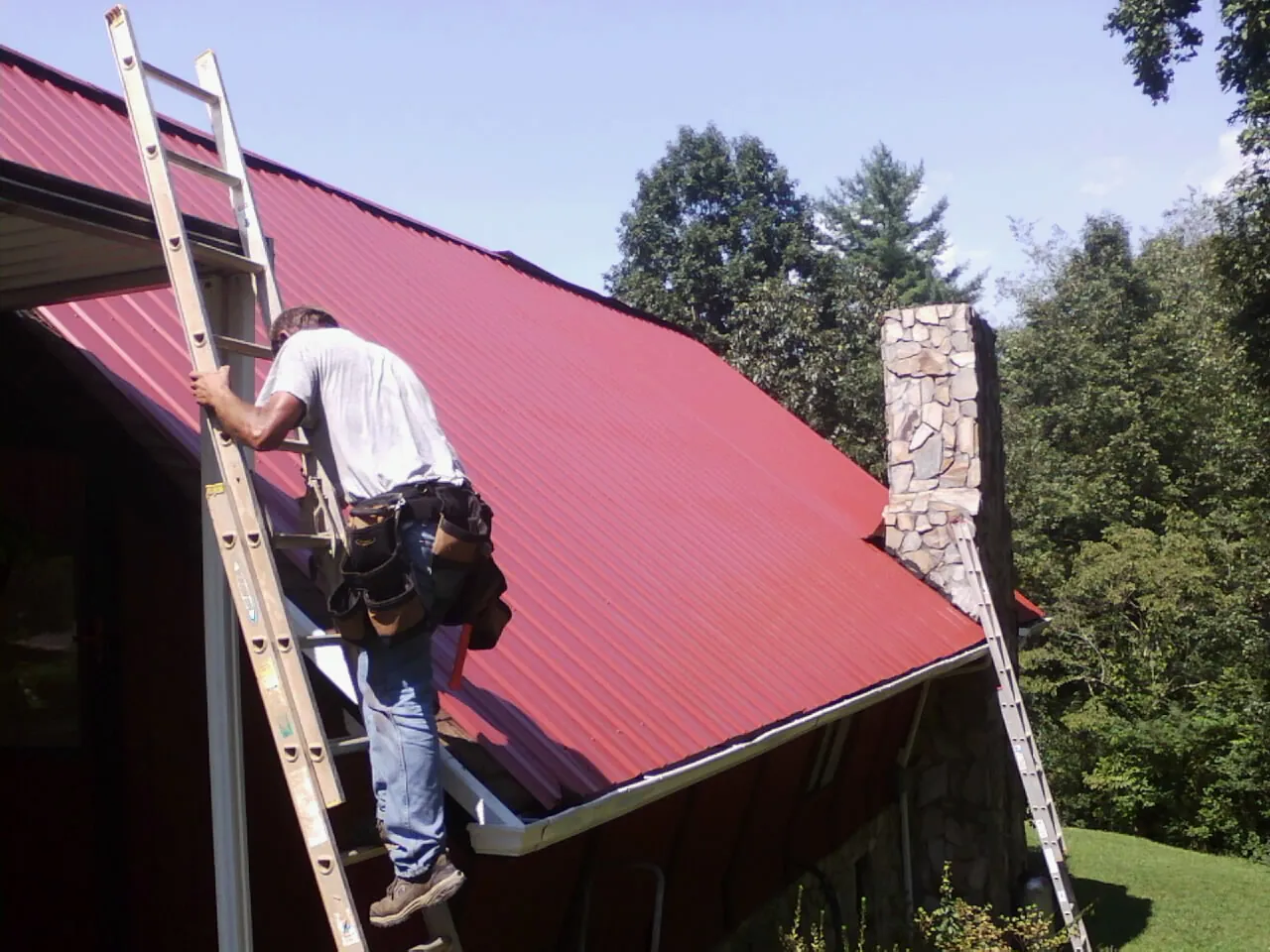 Roofer on a ladder beside a red metal roof and stone chimney — Soffit & Fascia Repair by Roofing Brothers in Cathedral City
